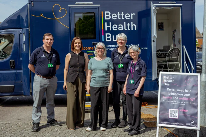 Two Settle colleagues stand with three partners from Hertfordshire County Council in front of the navy blue Hertfordshire Community Trust Better Health Bus, a mobile health service. A signboard next to the group provides information about the service.