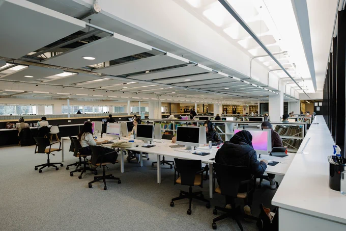 Photo taken from Zone D, level 03 in the LSBU Hub Library. Students sat at desks with Macbook computers on them and the bookshelves from Zone E can be seen in the background.