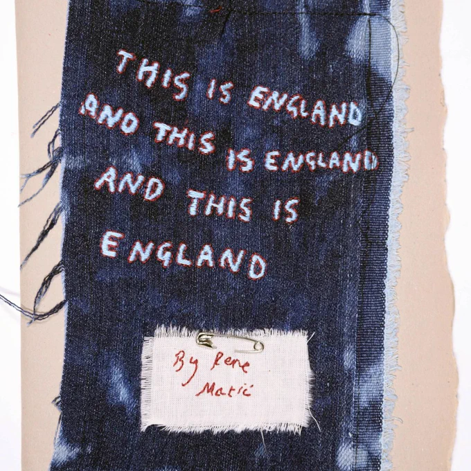 Embroidered denim artwork dissertaion cover – A piece of dark blue denim with white and red embroidered text reading "THIS IS ENGLAND AND THIS IS ENGLAND AND THIS IS ENGLAND", with a fabric label signed "By Rene Matić".