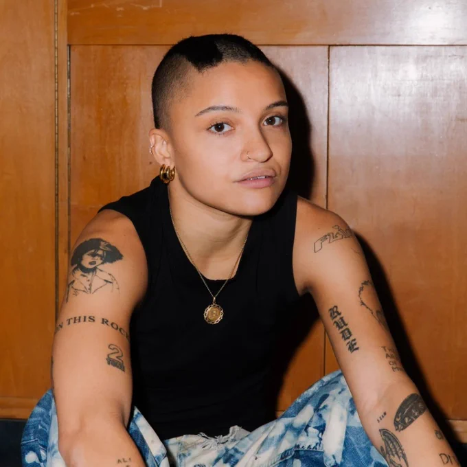Portrait of Rene Matić – Rene Matić sits in front of a wooden panelled wall, wearing a black vest top and patterned jeans, with tattoos visible on their arms