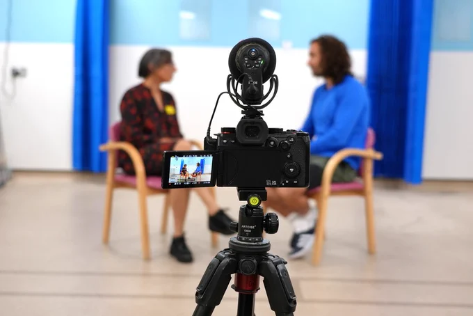 A video camera in the foreground is set up to film two people who are slightly blurred in the distance.