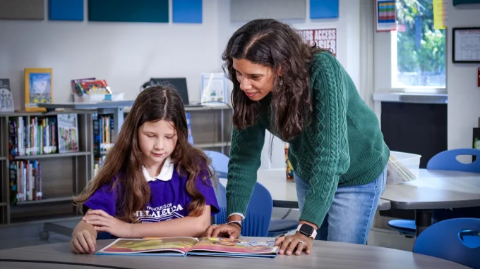 A teacher assists a student with reading a book at a table in a classroom.