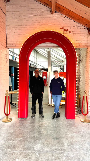 Two smiling members of the Slung Low team standing at the entrance, a lit up red velvet archway, to the space at the Warehouse