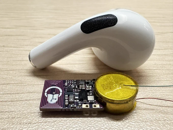 A compact custom PCB featuring a white logo of a tongue inside a mouth is connected to a small round yellow-laminated lithium battery. The board includes surface-mounted components and a visible coiled trace antenna. A white wireless earbud is placed next to it for scale comparison.