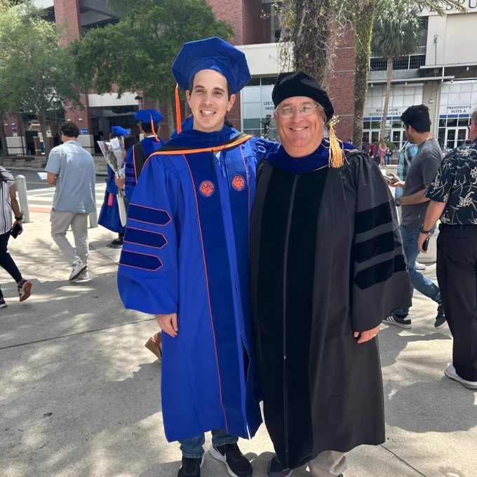 two people are standing outside wearing traditional university graduation robes, one in black and the other in dark blue