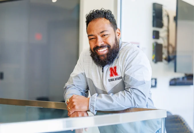 Ashley Wallace, global experiences coordinator in the Global Experiences Office, smiles as he leans over a rail. He is wearing a two-toned gray pullover with a red Nebraska block N and the words "Global Affairs."