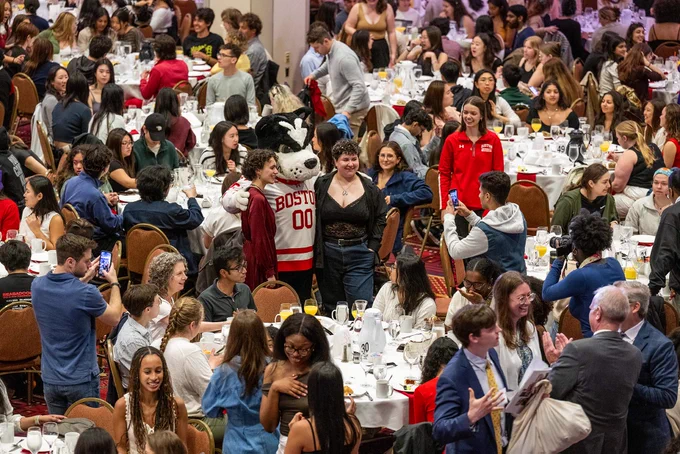 Photo: A large banquet hall filled with numerous round tables, each set with white tablecloths, plates, glasses, and cutlery. The room is crowded with people sitting at the tables and standing around. In the center of the image, a person in a Boston University mascot costume (a dog) wearing a red and white jersey with "BOSTON 00" on it is posing for photos with attendees. Many people are taking pictures or looking at their phones. The atmosphere appears lively and social.