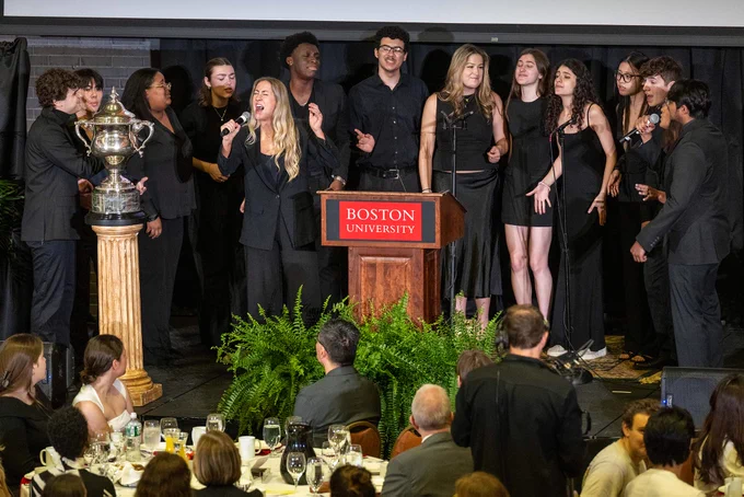 Photo: A group of people dressed in black are standing on a stage behind a podium with a "Boston University" sign. There is a large trophy on the left side of the stage. The audience is seated at tables with white tablecloths, glassware, and plates.