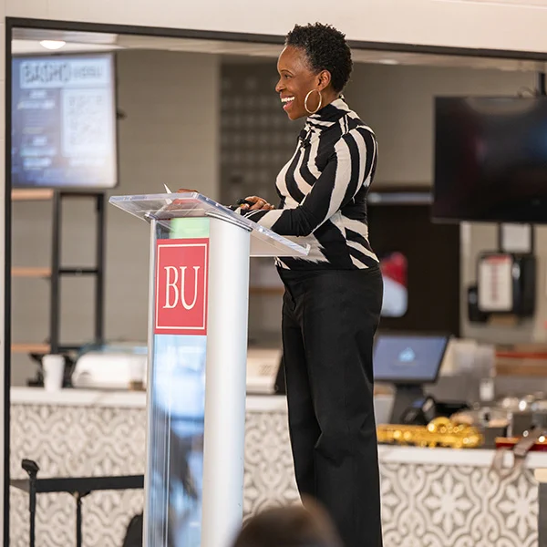 Photo: Boston University President Melissa Gilliam is standing at a podium with a "BU" logo on it, giving a presentation or speech. The person is wearing a black and white striped top and black pants. In the background, there are screens displaying information and various objects such as food trays and kitchen equipment. The setting appears to be an indoor event space or cafeteria.