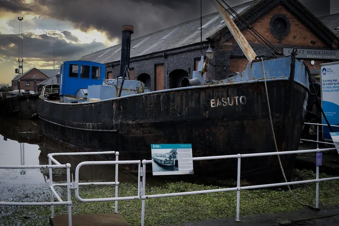 The preserved steam tug Basuto rests in a canal basin, surrounded by railings and signage, with stormy clouds overhead and historic warehouses in the background.