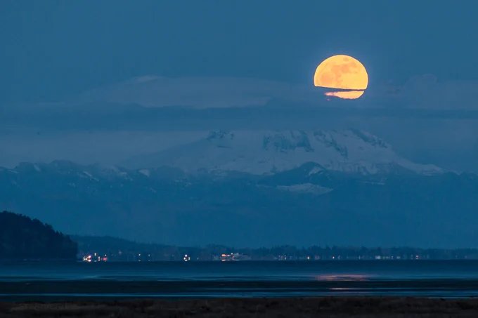 👨🏼‍🍳 Baker's Surprise 🗻🌝 The Full Pink Moon rises over the "Sleeping Giant" (aka Mt. Baker), the International Border of Canada/U.S.A. and the Bordertown of Blaine in Washington State 🇺🇸 

Looking southeast over Boundary Bay, the tip of Ocean Park in South Surrey on the left (with the White Rock Pier hiding behind it), over Semiahmoo Bay, to the shoreline of Blaine (20km/13mi away from where I stood), US Customs and Border Protection building on the left and the town of Blaine & Blaine Harbor Boating Center on the right, then beyond, over Lynden, WA to the majestic snow-capped Mt. Baker, 93km/58mi away.

Did you know that Mt. Baker is a glacier covered andestic stratovolcano? The last time it erupted was in 1880. It still shows signs of activity with steam plumes rising from Sherman Crater on cold clear days. It remains a potential hazard. 😳

Captured at blue hour near Tsawwassen, Delta, British Columbia, Canada 🇨🇦 ~ April 12, 2025 🌝