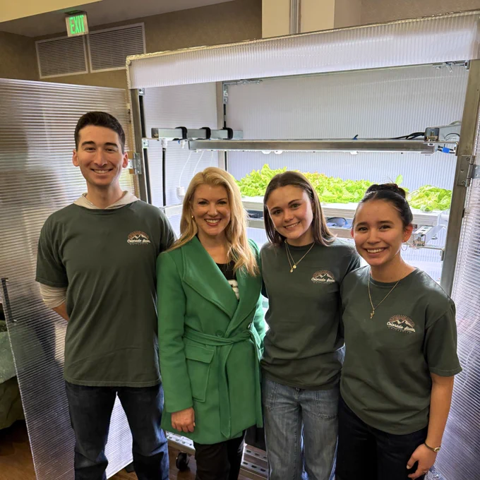 Four people stand smiling in front of an indoor greenhouse system with leafy greens growing under lights. Three students from CSU pose alongside President Amy Parsons.