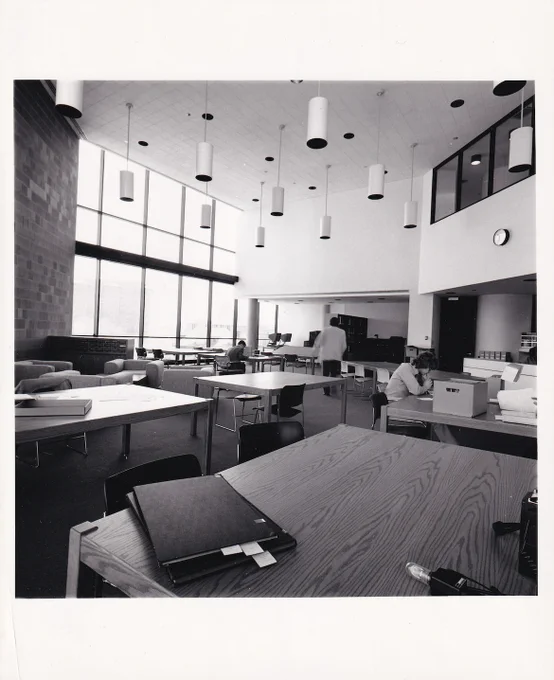 The Reading Room at the Reuther in 1975 in black and white. Large wooden tables hold archival material and boxes for researchers, huge windows let in natural light, and large lights hang from the ceiling above. 