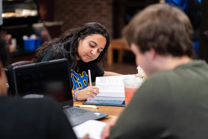 A student wearing a "Pitt" t-shirt writes in their notebook while sitting at a table with others in Hanley Library.