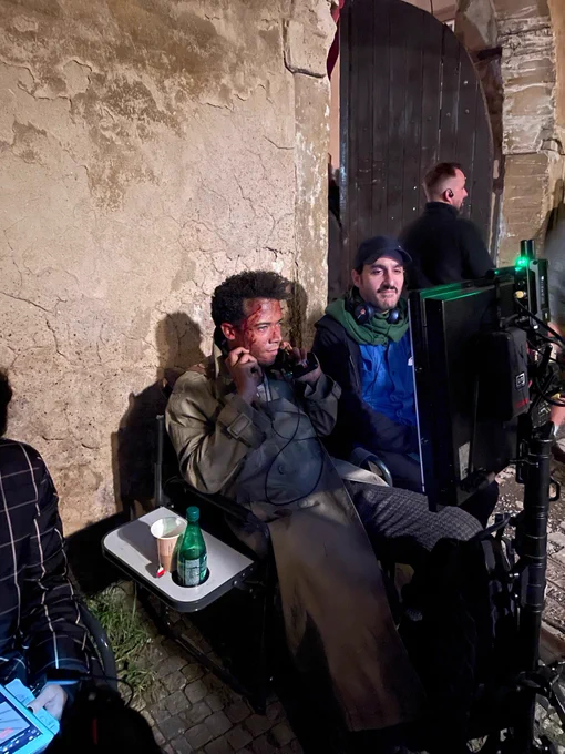Jacob Anderson, in full Louis makeup and costume, sits in front of the camera monitor, next to Director Levan Akin. Jacob is holding the headphones under his ears, expressionless, as he watches the scene being filmed. 