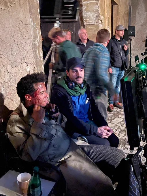 Shot more from that side, Jacob Anderson, in full Louis makeup and costume, sits in front of the camera monitor, next to Director Levan Akin. Jacob is holding the headphones under his ears, expressionless, watching the scene being filmed. 