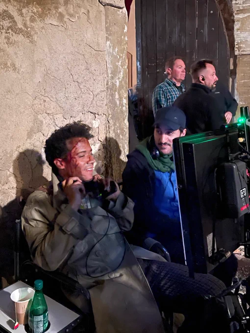 Jacob Anderson, in full Louis makeup and costume, sits in front of the camera monitor, next to Director Levan Akin. Jacob is holding the headphones under his ears, smiling as he watches the scene being filmed. 