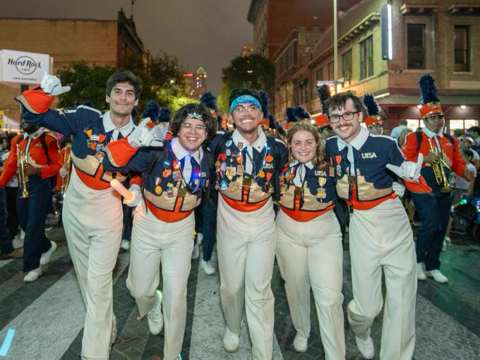 A group of five University of Texas at San Antonio marching band members posing joyfully, wearing uniforms adorned with medals, during an outdoor event at night.