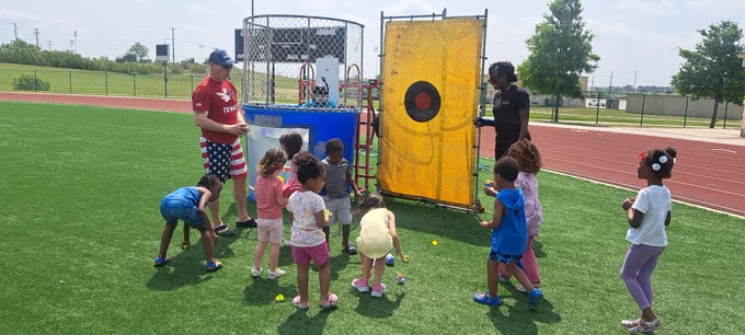 Command Sgt. Maj. Loyd Rhoades, U.S. Army Garrison-Fort Cavazos command sergeant major, tosses a ball to dunk Greta Buccellato, Fort Cavazos deputy to the garrison commander, with the help of a group of children during the 20th Annual Garrison Org Day at Fort Cavazos.