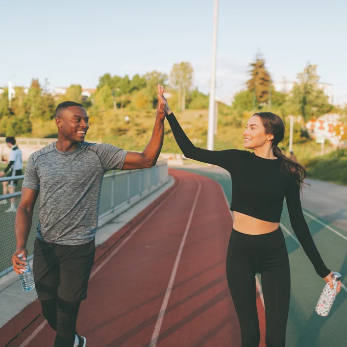 Two people high-five while walking on a running track, both holding water bottles and smiling after a workout.