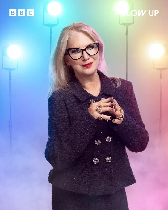 Judge of Glow Up and make-up artist Val Garland stands in front of a pastel rainbow background. She has black rimmed glasses and has her hands clasped in front of her. She has rings on each finger. She wears a purple tweed suit that has a long matching skirt.