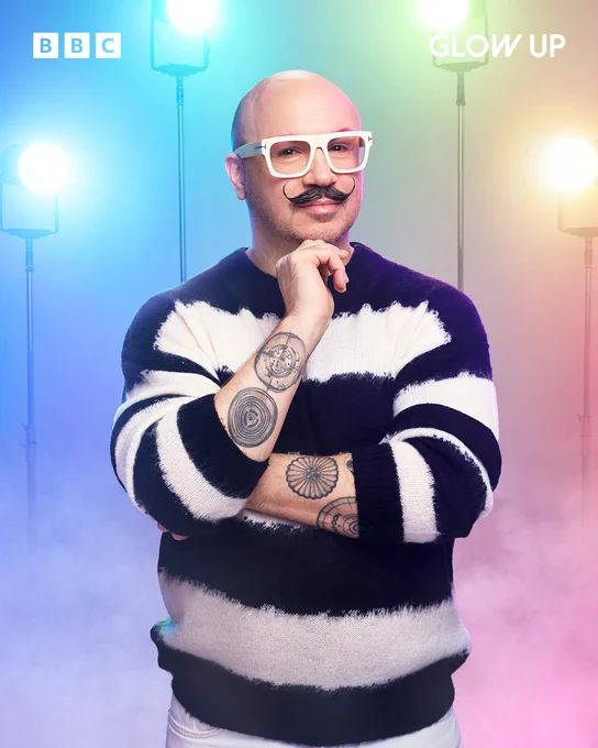 Judge of Glow Up and make-up artist Dom Skinner stands in front of a pastel rainbow background. He has a handle bar moustache and wears white thick rimmed glasses. He has circular tattoos on both arms and his sleeves are pushed up to his elbows. He wears a white and blue stripped long sleeve t-shirt. 
