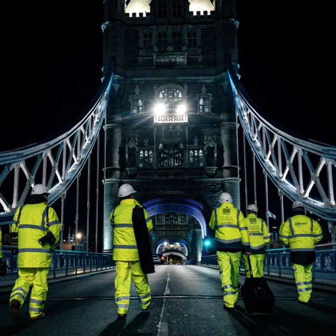 Six people wearing high visibility PPE with white helmets walk across a silent Tower Bridge at night.