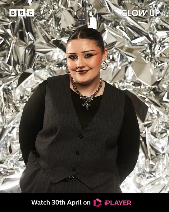 Ailish stands in front of a shiny background crinkled like foil. She wears a black oinstripe waistcoat with a black long sleeve shirt underneath. She has a slicked bun with dark hair and a bold make-up look. She has a silver chain pendant on. 