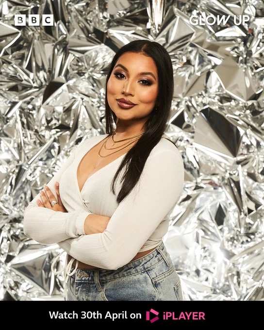 Jade stands facing the left with her body turned towards the camera. She wears a white long sleeve top  and has her arms crossed. She has three gold chains on and a bold face of makeup. 