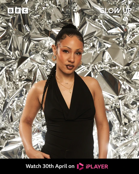 Cherise stands in front of a shiny background crinkled like foil, she wears a black v necked top with no sleeves and a gold cross chain. She has a braided ponytail down her back. 