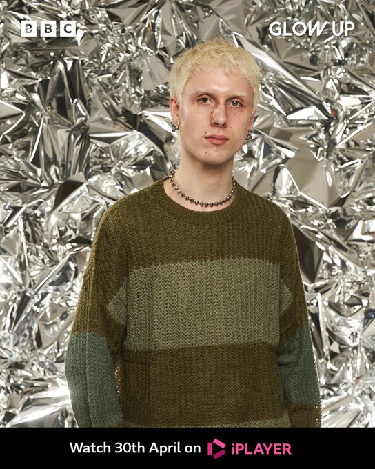 Jake stands in front of a shiny background. He has bleach blonde hair and matching eyebrows. He has a tanned face and dark brown freckles. He wears a green wide stripped knit jumper, which has different shades of green. 