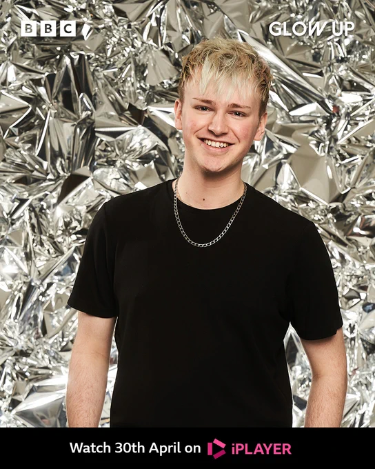 Jamie stands in front of a shiny foil background. He has dark hair on the sides which is long on the front with blonde highlights. He wears a silver long chain and a plain black t-shirt. 