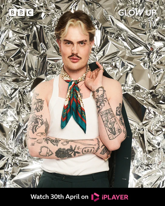 Joshua stands in front of a shiny foil-like backdrop. He has short dark hair which sits above his ears, with blonde curtain bangs. He also had dark brown eyebrows wirh a slit in the right one. He wears a silk scarf knotted at his neck and has bare arms which are covered in tattoos on his forearms. He has a drak blazer over his right shoulder which he holds with one hand. 