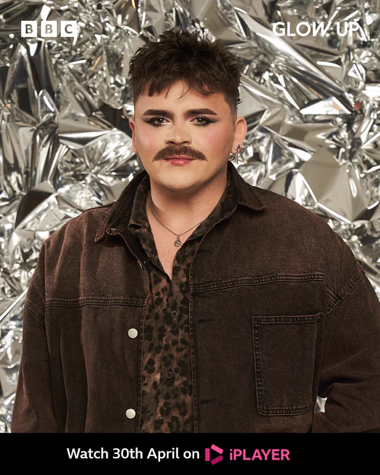 Joe stands in front of a shiny foil-like backdrop. He has messy dark brown hair and matching straight brows and a handle-bar moustache. He wears a cheetah print button up shirt and over the top a brown suede style jacket. 