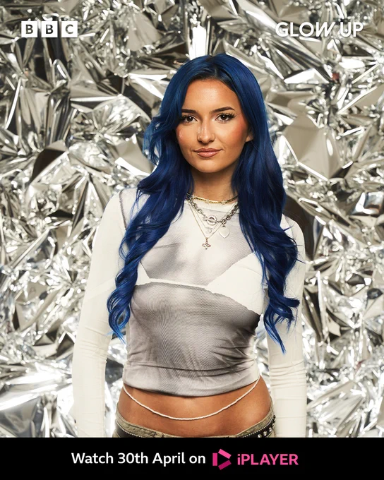 Mia stands in front of a shiny foil-like backdrop. She has long blue hair which is curled and wears a long sleeved white top with a bra imprint over the top. She wears a white small waist bead and low-rise jeans. 