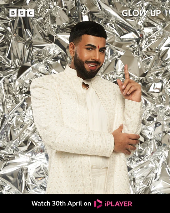 Rahual stands in front of a shiny foil-like backdrop. He has black hair with a clean line-up and has a traditional red bindi in the middle of his forehead. He wears a white embroidered Indian suit with long sleeves. He rests his left hand on his right elbow with his arm streched upwards. 