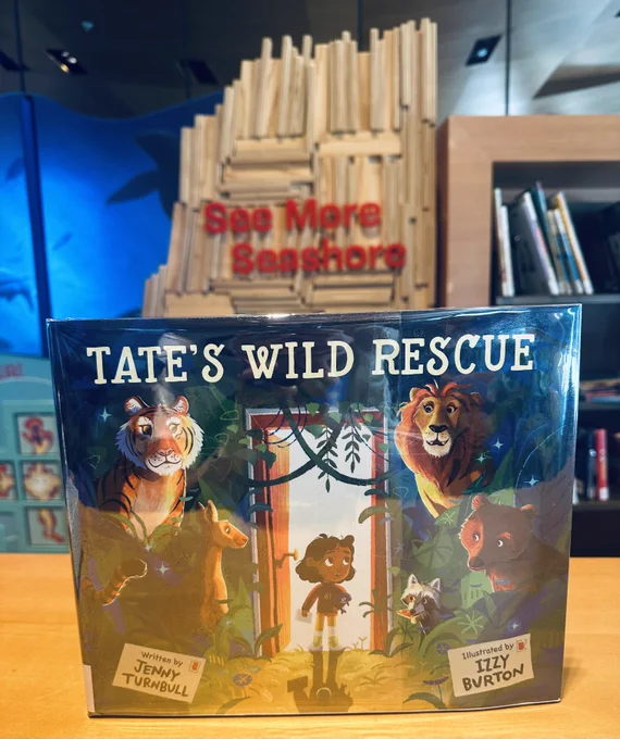 The children's book Tate’s Wild Rescue is displayed upright on a wooden surface in a library. The book cover features a young girl standing at the entrance of a forest, surrounded by illustrated animals including a tiger, kangaroo, lion, raccoon, and bear. The title is written in bold white letters at the top. The book is written by Jenny Turnbull and illustrated by Izzy Burton. In the background, there is a wooden art installation with the words "See More Seashore" in red letters, and bookshelves filled with books.