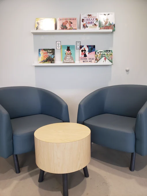 Two blue chairs sit opposite each other at a low circular side table. On the wall are two rows of shelving with picture books shelved outward on them. 