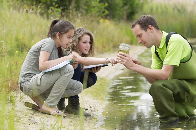 Learners and an educator outdoors by the side of a pond. The educator is holding a glass jar of pond water as the students record observations in their notebooks.