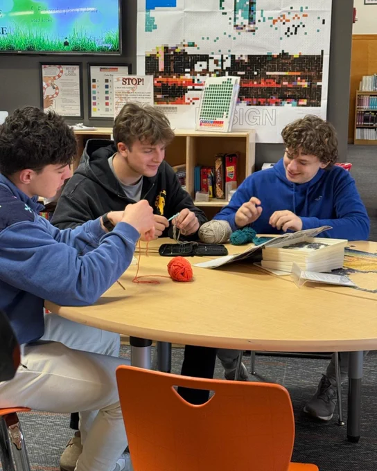 Three students sit at library table while crocheting.
