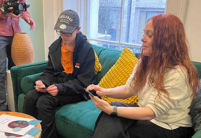 A young person sitting on a sofa, with an adult on the right and a camera to the left.