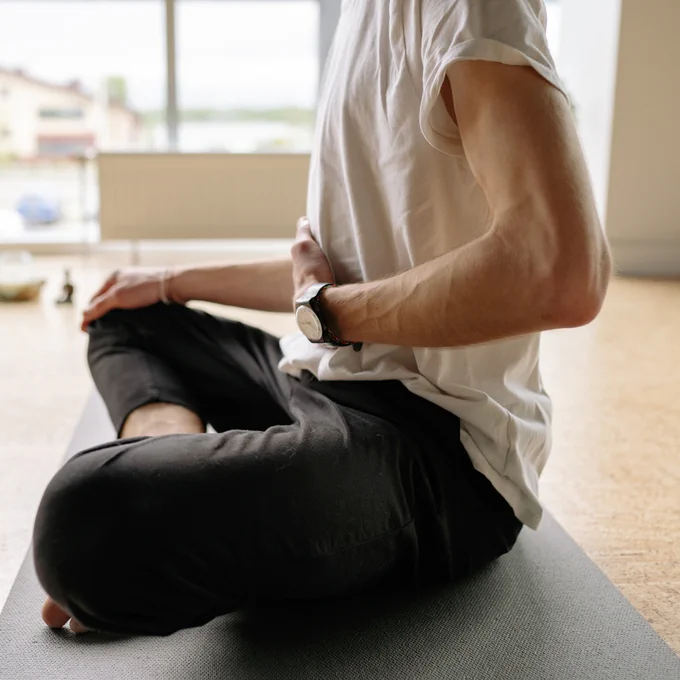 A person sits cross-legged on a yoga mat in a bright room, resting one hand on their stomach while practicing deep breathing.