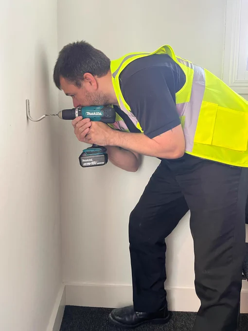 Direct Access accessible media lead Craig Acton drilling a hook into a wall in a hallway. He is wearing a green hi-vis workers jacket, black trousers and black shoes.