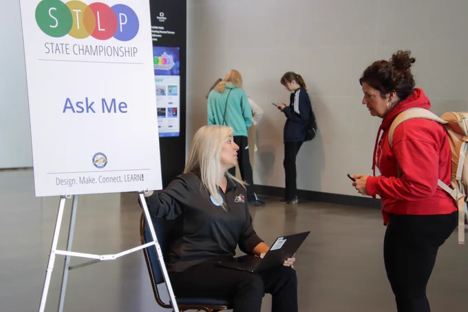 A woman talks to another woman by a sign that reads “Ask Me”