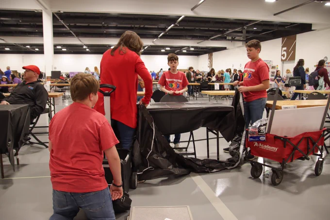 A woman and three students put a black table cloth over a table as a cart with more items from their STLP exhibit sits off to the side