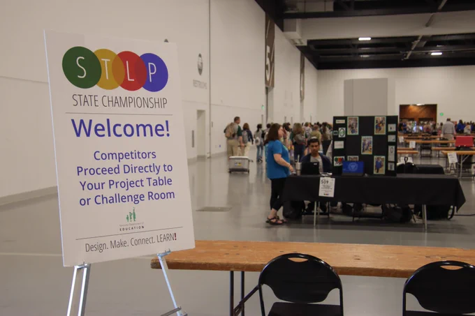 A sign reads “STLP State Championship. Welcome! Competitors proceed directly to your project table or challenge room” as students prepare their exhibits in the background