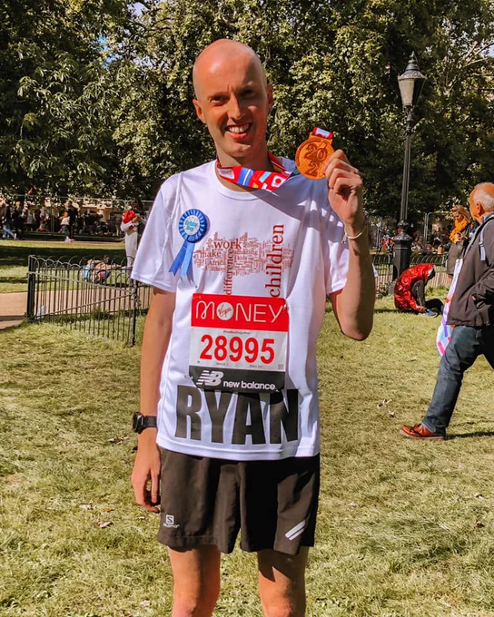 A smiling man in a white African Children's Fund t-shirt with RYAN written in big letters on the front, below his London Marathon race number. He is wearing a London Marathon medal around his neck, which he is holding up to the camera.