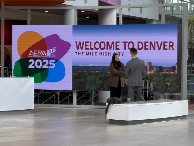 Two people stand in front of an electronic sign that says Welcome to Denver with the AERA 2025 Annual Meeting logo