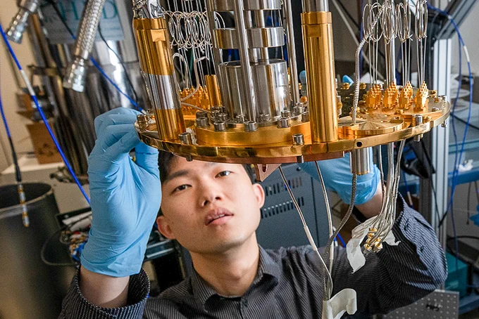 A researcher in a lab crouched below a silver and gold quantum computer. They are making adjustments to the bottom edge.