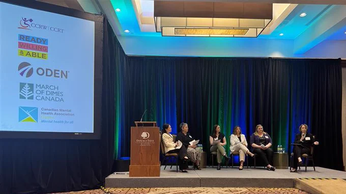 In a conference setting, six women are seated on a low stage, participating in a panel discussion. To the left is a large screen with the following logos: CCRW; Ready, Willing & Able; ODEN; March of Dimes Canada; Canadian Mental Health Association.